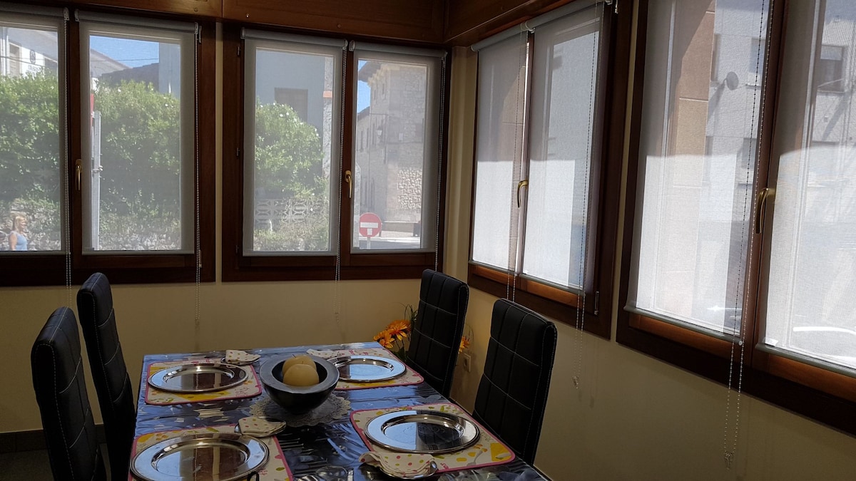 A dining area is positioned near large windows that allow natural light to fill the space. A colorful tablecloth covers the table, set for six with plates and utensils arranged neatly. The windows feature sheer coverings, providing a gentle diffusion of light.
