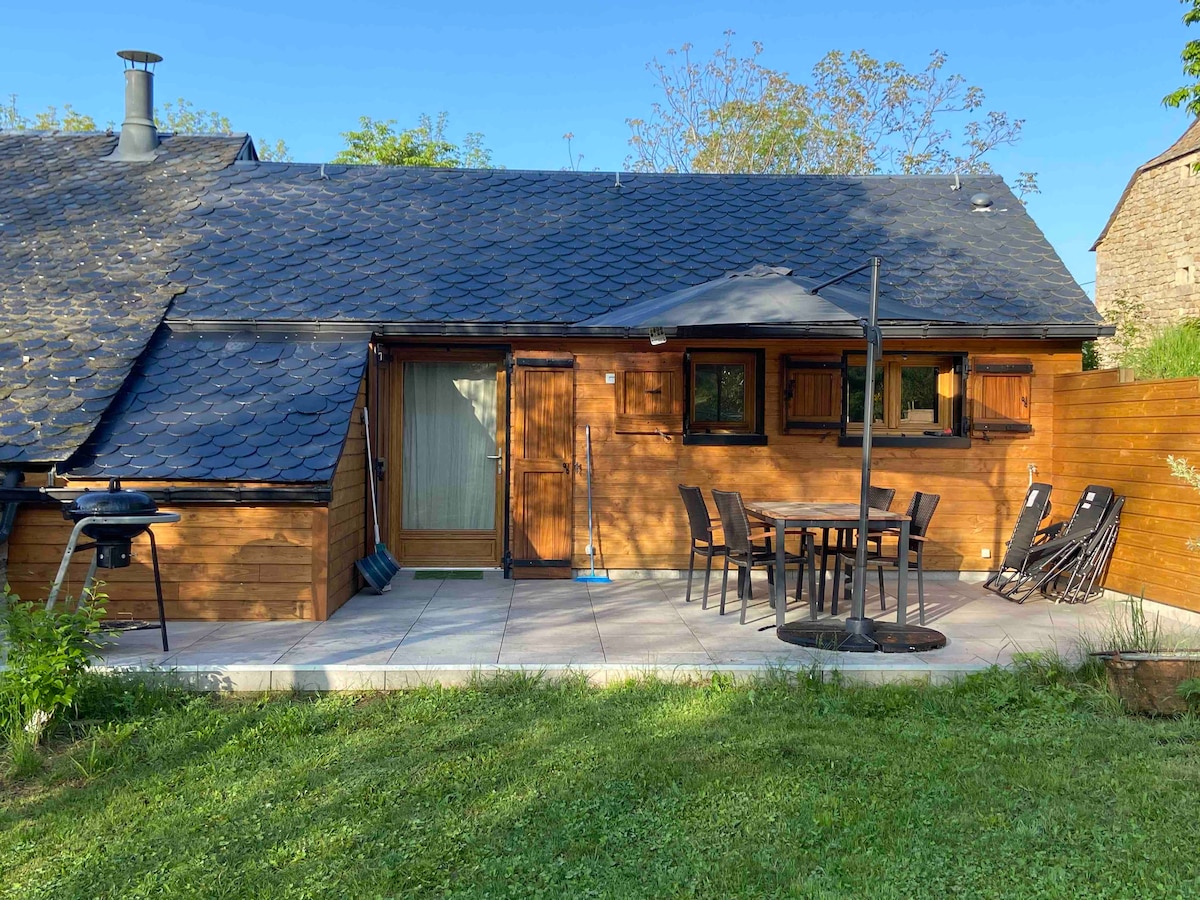 A wooden exterior is featured in this single-story accommodation, highlighted by a sloped roof and large windows. An outdoor dining area with a round table and chairs is visible on a tiled patio, complemented by a barbecue grill and lounge chairs on a grassy lawn.