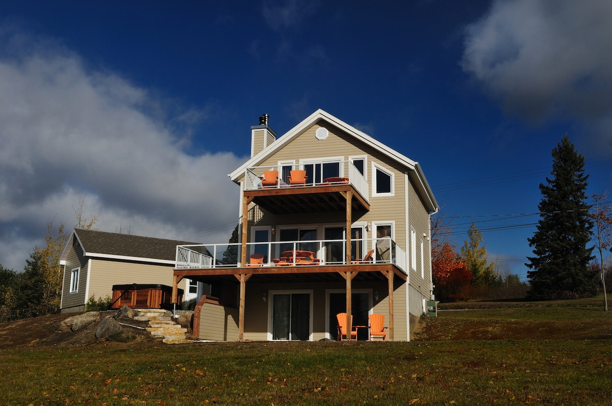 The residence features a three-story structure with a spacious deck adorned with orange chairs. A hot tub is located on the lower level, surrounded by natural landscaping. Large glass doors provide access to the interior, while the sky showcases a mix of clouds and blue tones.