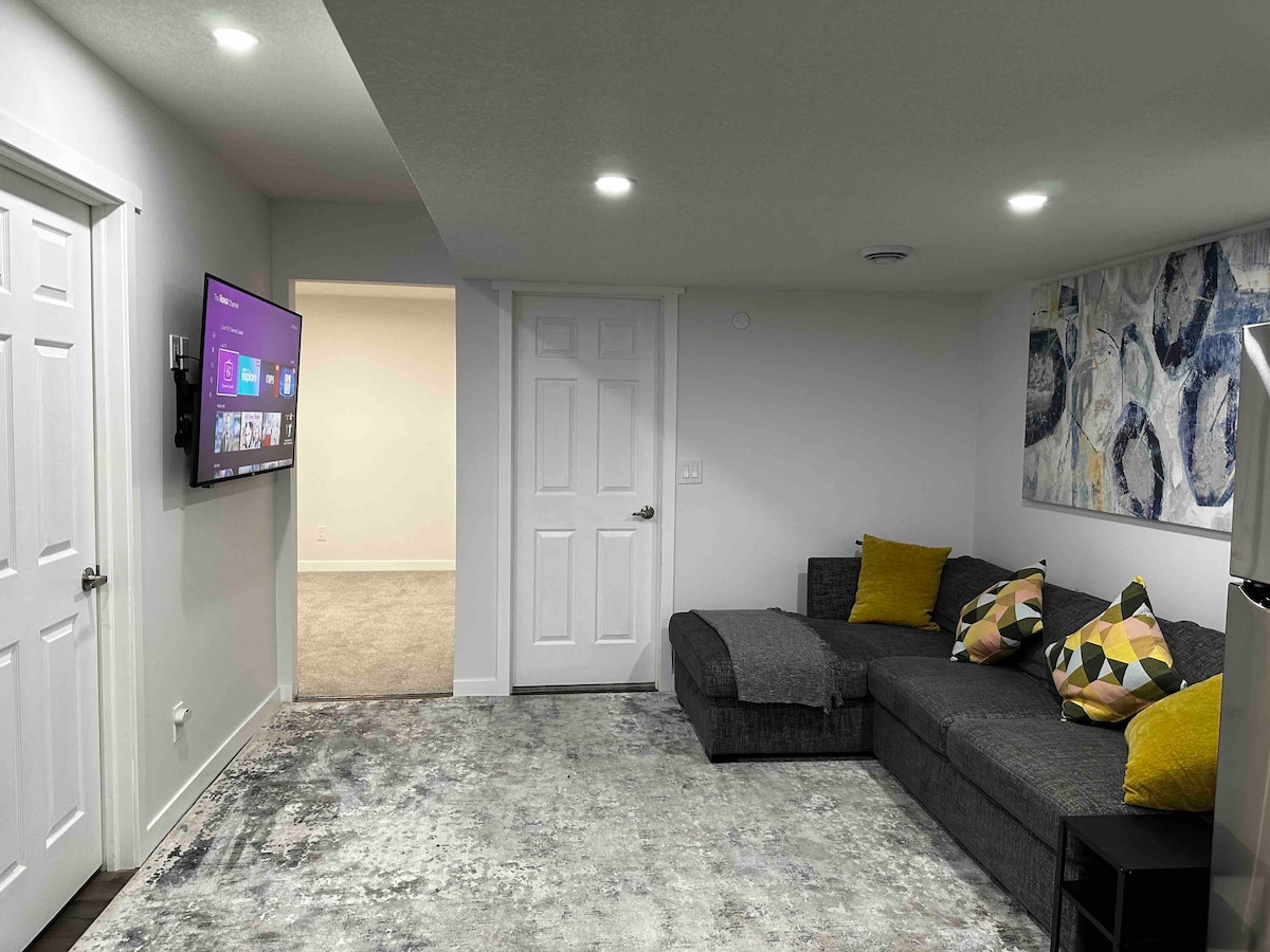 A modern living area is presented with a comfortable sectional sofa adorned with decorative cushions. A wall-mounted television is visible across from the seating space, with soft, recessed lighting illuminating the room. An abstract wall art piece in cool tones enhances the ambiance.