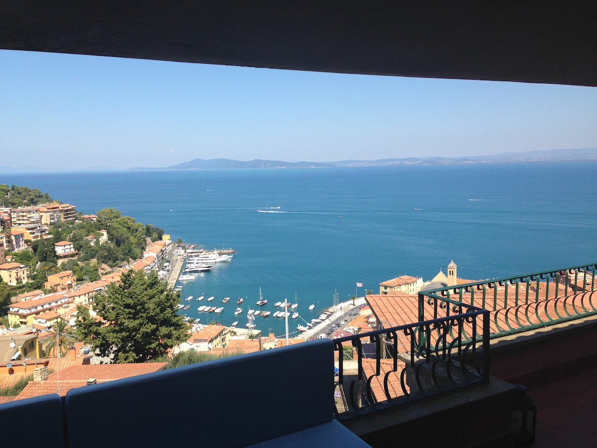 A scenic view of the coastline is displayed, showcasing a vibrant blue sea dotted with boats. The image captures the distant hills on the horizon, framed by the architecture of the nearby buildings, and offers a glimpse of the bright sky above.