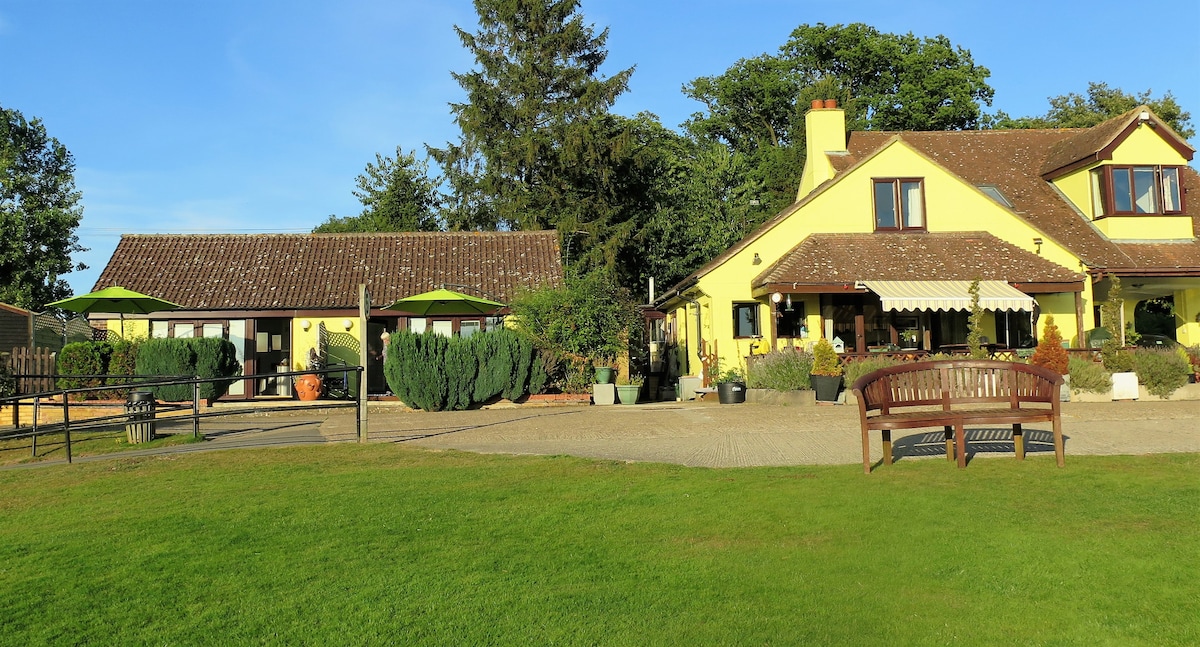 A yellow house with a sloping roof and dark trim features a welcoming porch. Adjacent, a separate building with a tiled roof is visible. Lush greenery surrounds the area, along with neatly placed potted plants and a wooden bench on a grassy lawn.
