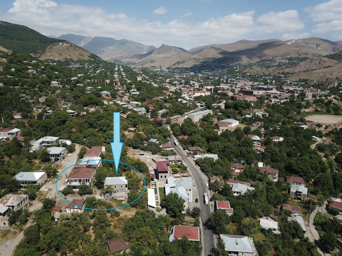 An aerial view captures the guest house amidst a green, hilly landscape in Goris, Armenia. Surrounding buildings can be seen, with mountains in the background and a clear blue sky above. The property is highlighted, showcasing its location within the community.