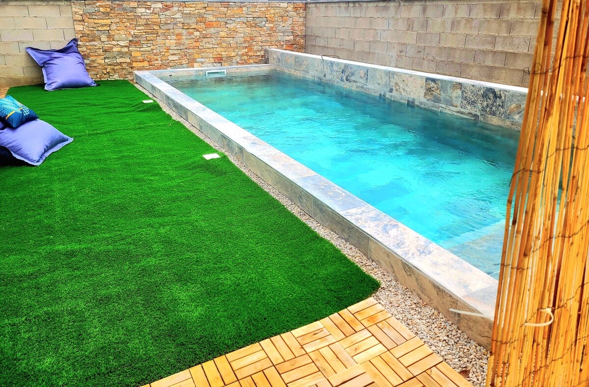 A shallow pool is surrounded by artificial grass, creating a lush green area for relaxation. Colorful cushions are placed nearby for comfort. The pool's clear blue water invites refreshing moments, while stone walls provide privacy and a natural aesthetic.