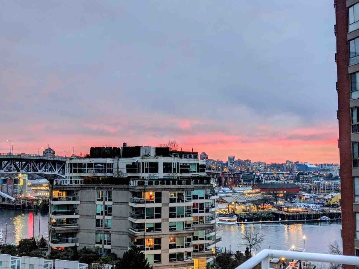A vibrant sunset is showcased over the city skyline, with soft pink and purple hues illuminating the clouds. The waterfront area and nearby buildings are visible in the foreground, with the bridge and distant structures framed against the colorful sky.