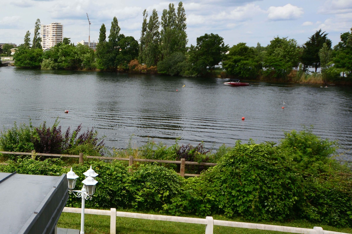 A serene view of calm water reflects the greenery lining the shore. Lush trees provide a natural backdrop, while a small boat is visible on the water. Soft ripples and distant buildings create a peaceful atmosphere by the water's edge.