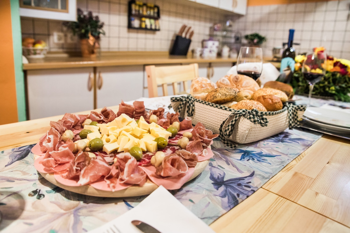 A wooden table is set with a round platter featuring an assortment of cured meats, cheese, and olives, complemented by a basket of fresh rolls. The kitchen background displays cabinetry, a countertop, and greenery, contributing to a warm dining atmosphere.
