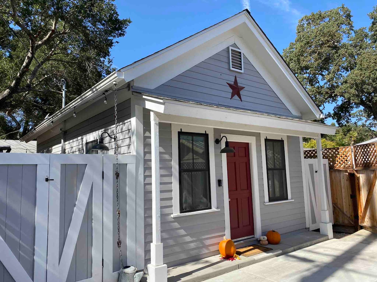 This charming cottage features light grey siding with white trim and a welcoming red front door. A star decoration hangs above the door, and two orange pumpkins adorn the entryway. The surrounding area includes a wooden fence and vibrant greenery from nearby trees.