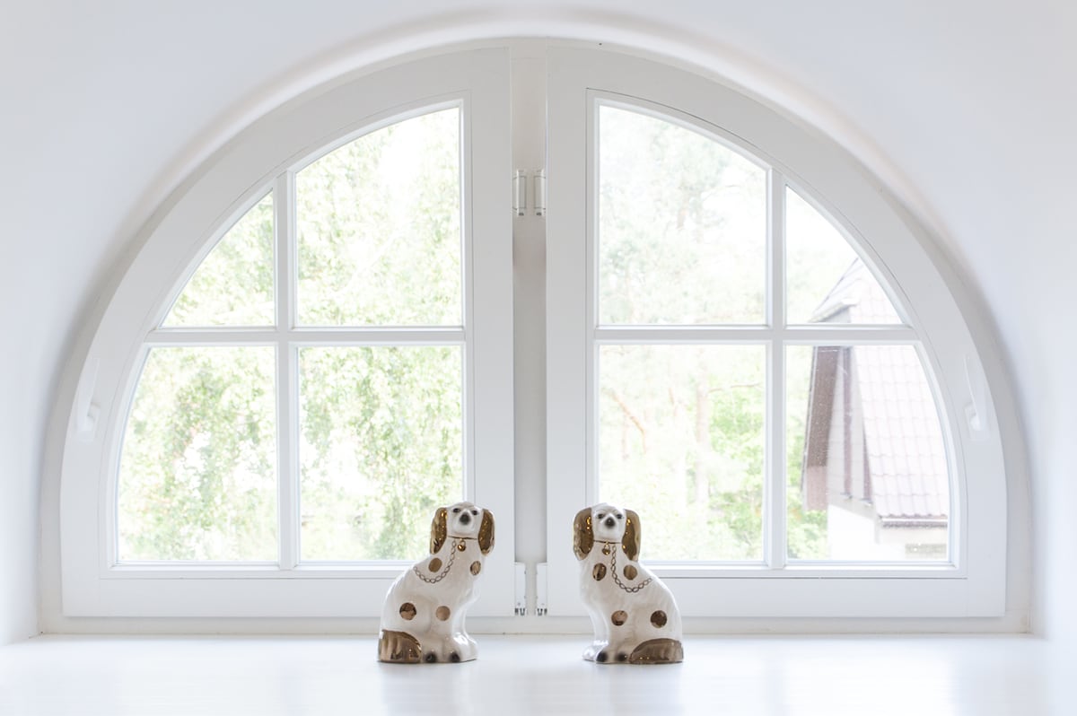 Two ceramic dog figurines are placed on a white surface beneath a large arched window. The window features multiple panes, allowing natural light to illuminate the space and providing a view of greenery outside.