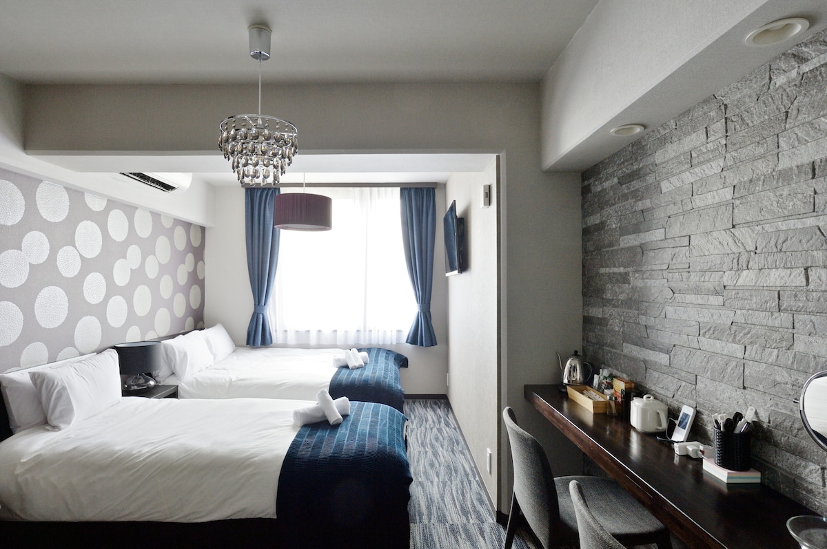 The room features two spacious beds dressed in white bedding, complemented by blue accents. A stone accent wall provides a modern touch. Natural light fills the space through a large window, which is framed by blue curtains. A work desk with chairs and a TV is also present.