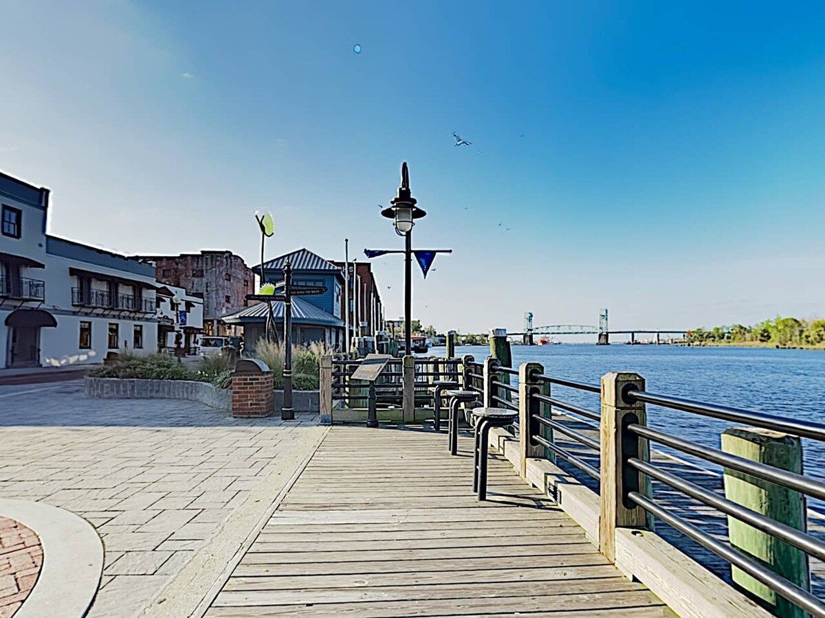 A scenic waterfront walkway is presented, lined with wooden planks and a black railing. A few benches provide seating along the path, which is accompanied by a clear view of the water. Buildings and greenery are visible in the background, along with a bridge in the distance.