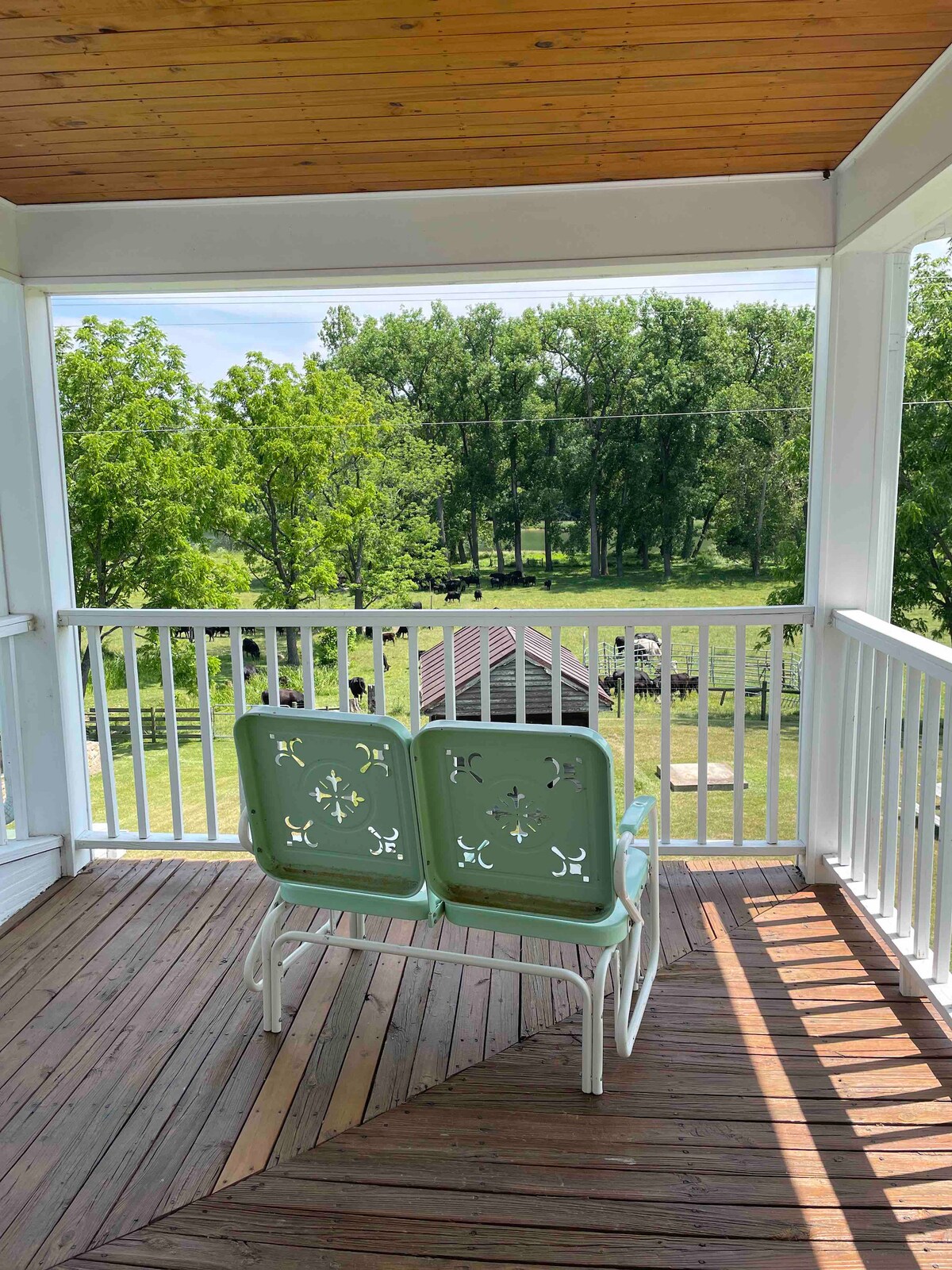 A covered porch features two green chairs positioned for enjoying views of the surrounding greenery and distant barn. Sunlight filters through the trees, creating patterns on the wooden porch floor.