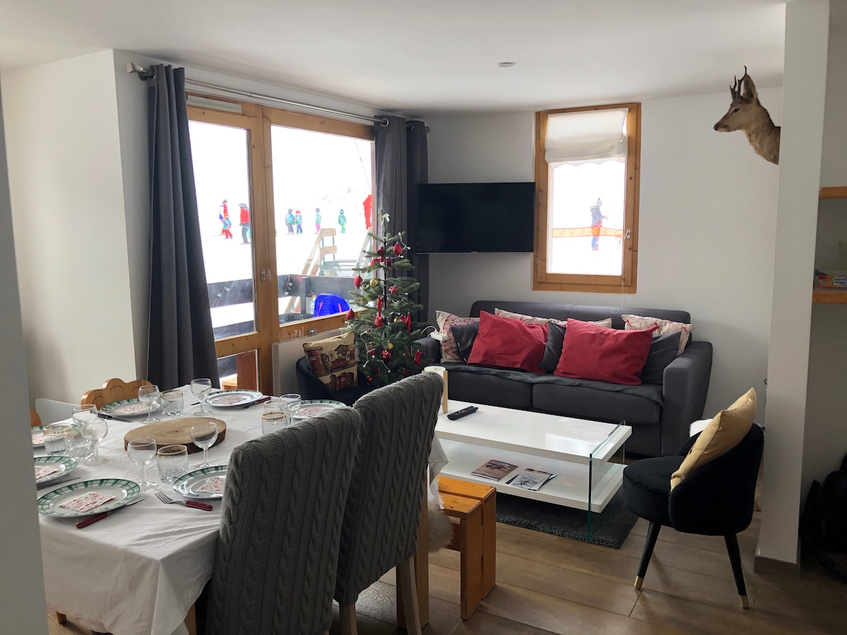 A bright living area features large windows overlooking a snowy landscape. A modern sofa adorned with cushions sits alongside a glass coffee table. A dining table is set with plates, and a small Christmas tree adds a festive touch. A television is mounted on the wall.