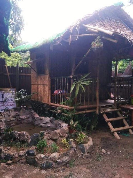 A traditional Bahay Kubo nipa hut stands amid lush greenery, featuring a thatched roof and bamboo construction. A small garden with stones surrounds the base, while a sign indicating Antipolo City can be seen nearby. The structure integrates with the natural environment.