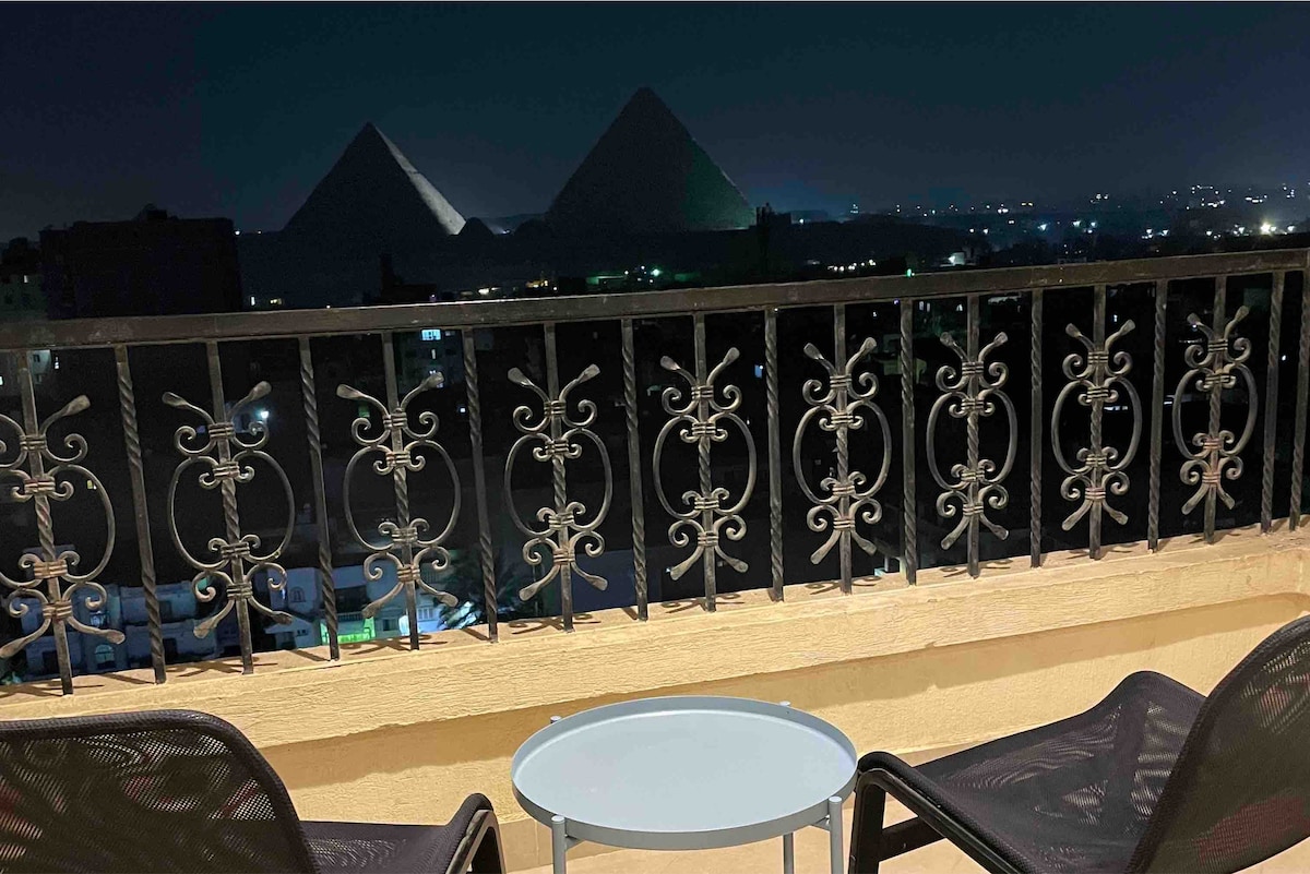 A balcony chairs are positioned around a small round table, offering unobstructed views of the Great Pyramids at night. The silhouette of the pyramids is contrasted against the dark sky, enhancing the serene atmosphere of the outdoor space.