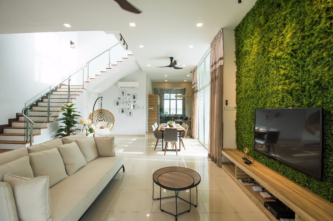 A spacious living area features a comfortable light-colored sofa facing a wall-mounted television. Lush green vertical gardens complement the sleek design. A large dining table is visible in the background, illuminated by natural light from the large windows.