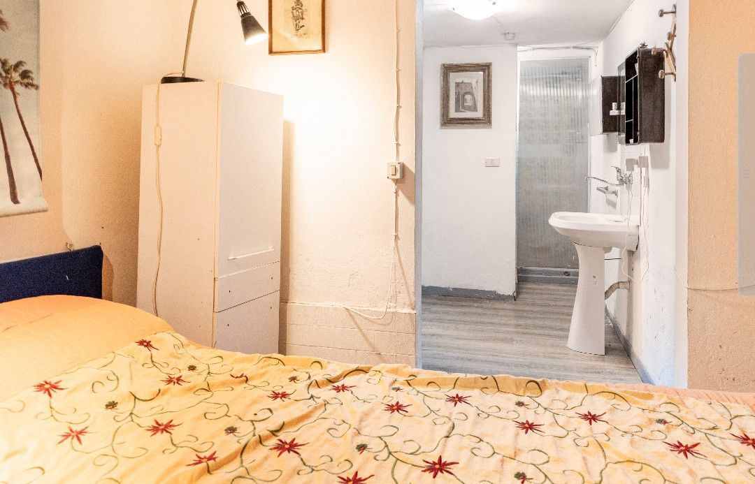 A cozy sleeping area is featured with a floral-patterned blanket on a spacious bed. The adjacent bathroom area is accessible, with modern fixtures and light-colored flooring. A small white cabinet and wall lamps provide illumination, creating a functional and inviting space.