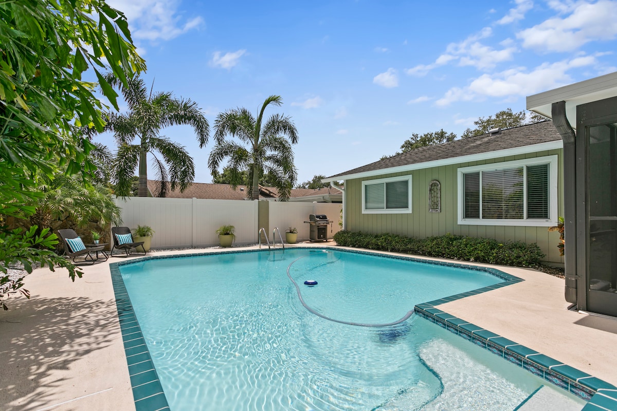 A tranquil private backyard features a clear pool surrounded by concrete decking and lush landscaping. Lounge chairs are positioned near the water, with palm trees providing shade. A charcoal grill is visible on the side, and the home’s exterior is painted in soft, inviting tones.