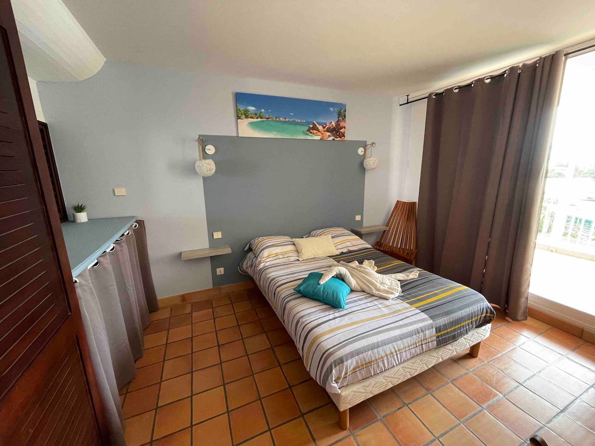 A sleeping area is presented with a striped bedspread laid over a spacious bed. Soft natural light filters through large windows covered by dark curtains. Decorative elements include two wall-mounted lamps and a beach-themed artwork above the bed, creating a relaxing ambiance.