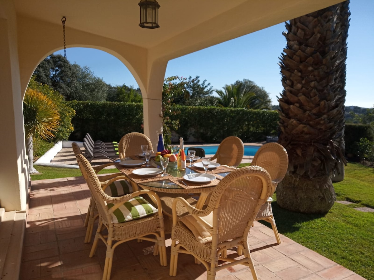 An outdoor dining area is set beneath an archway, featuring a round table surrounded by six wicker chairs. A vibrant table setting includes glasses and a vase with flowers. Lush greenery and a private pool can be seen in the background, enhancing the tranquil atmosphere.