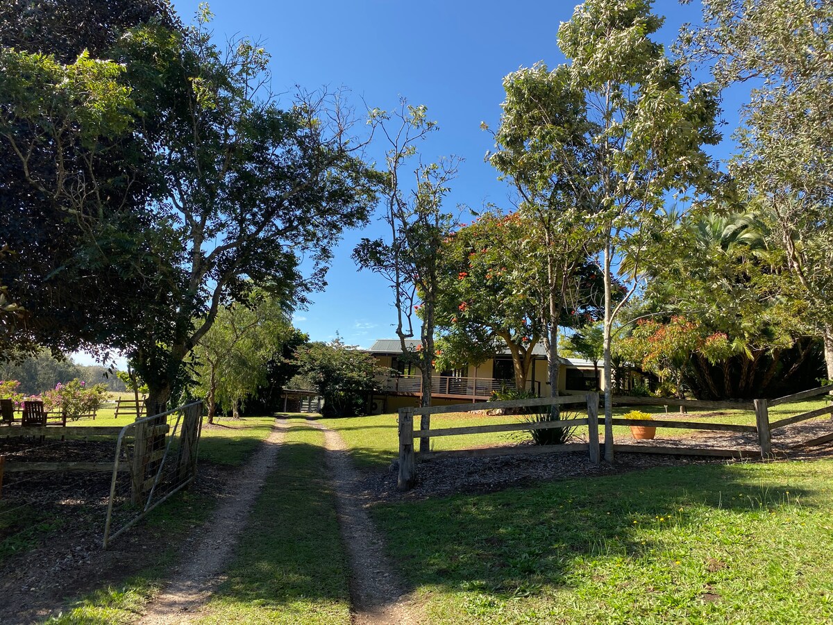 A gravel path leads through a lush green landscape, bordered by trees and shrubs. The farmhouse is visible in the background, surrounded by well-maintained gardens, inviting an atmosphere of tranquility and nature.
