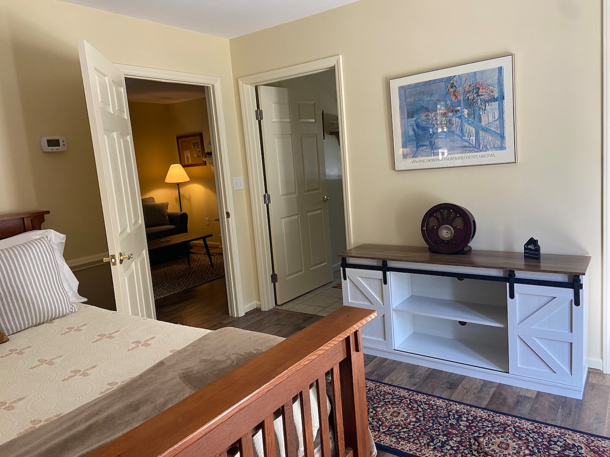 A bedroom area displays a full-sized bed with neutral bedding, complemented by a decorative bed frame. A stylish console table sits against the wall, featuring a decorative item. Doorways lead to the bathroom and living area, while soft lighting brightens the space.