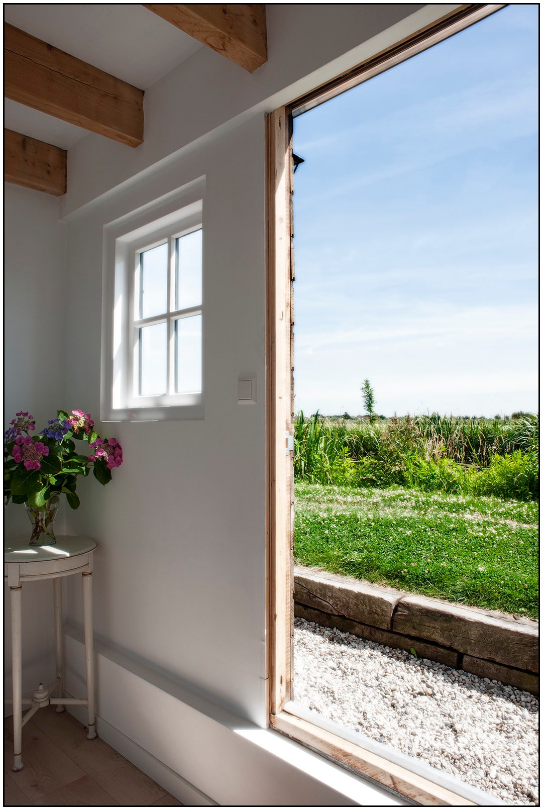 An open door reveals a bright view of lush greenery outside. A small table with a vase of flowers is positioned beside the door, and sunlight illuminates the interior space, enhancing the airy atmosphere.