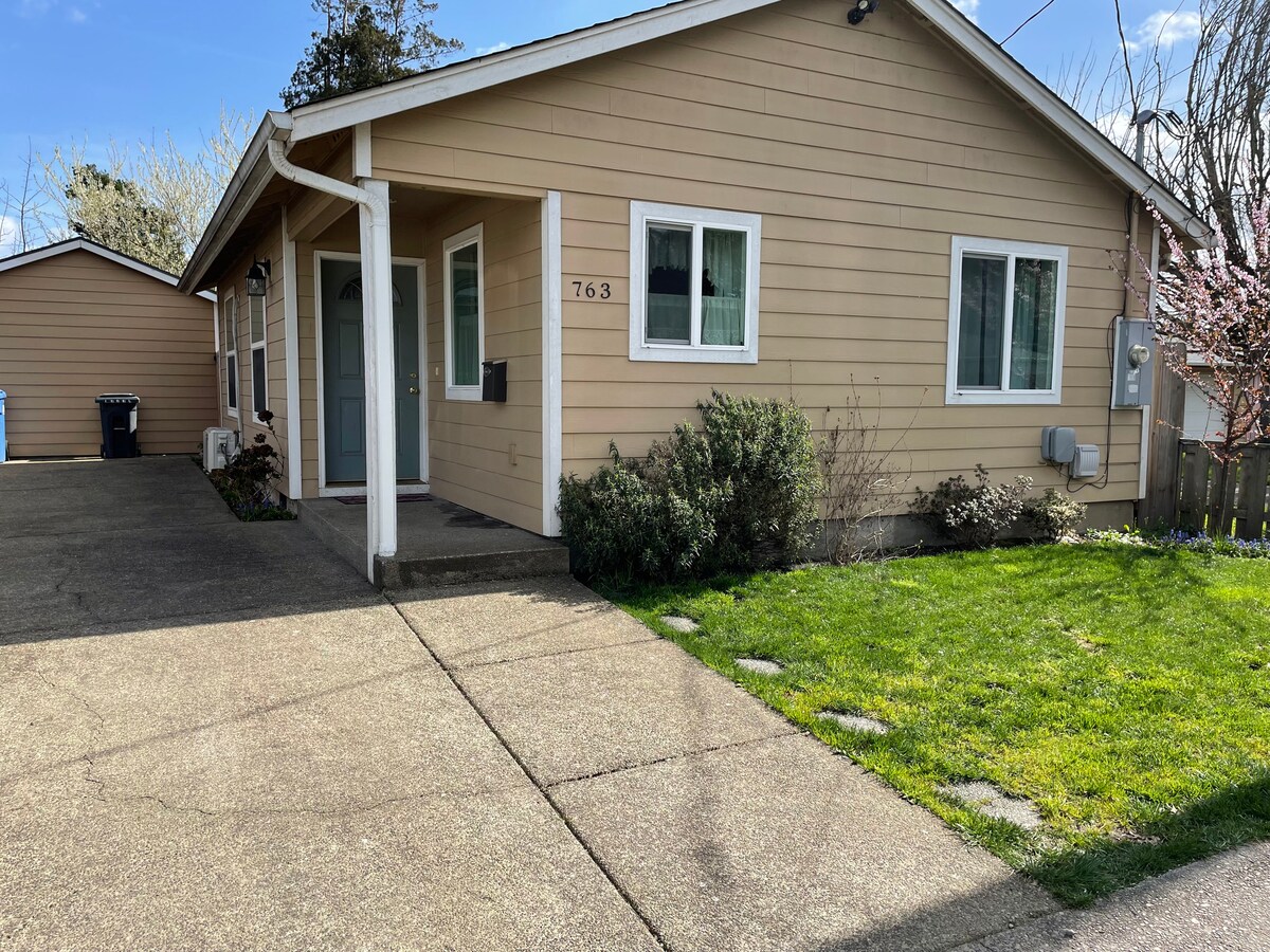 A single-story house with a light beige exterior is shown, accompanied by a well-maintained front yard featuring green grass and shrubs. Two large windows are visible on the front, along with a welcoming covered entryway. A driveway leads to an adjacent garage.