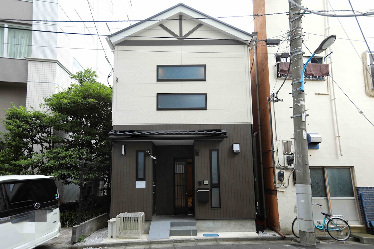 A modern two-story house is presented, featuring a neutral-colored facade with large windows. The entrance is marked by a simple door, flanked by a small porch area. The surrounding greenery provides a natural contrast to the building's contemporary design.