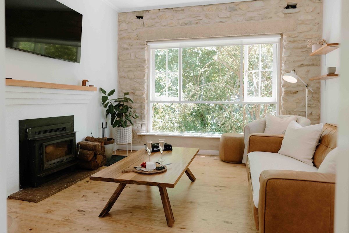 A living area features a stone wall complemented by a wood fireplace. A large window provides ample natural light, framing views of leafy trees outside. A comfortable sofa sits near a wooden coffee table, which is arranged with drinks and snacks, enhancing the inviting atmosphere.
