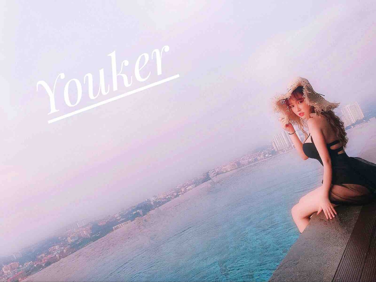 An infinity pool extends toward the horizon, providing a panoramic view of the cityscape. Soft pastel colors dominate the sky, blending with the water's edge. A figure is seated at the pool's perimeter, presenting a serene atmosphere and inviting setting.