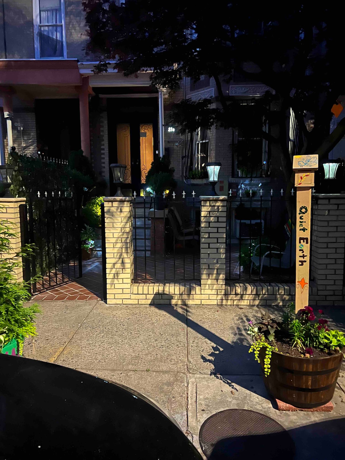 A welcoming entrance is framed by a low brick wall with a decorative sign reading 'Quiet Earth.' Lush greenery and potted plants are arranged alongside the pathway, while lighting fixtures add an inviting touch to the serene outdoor space.
