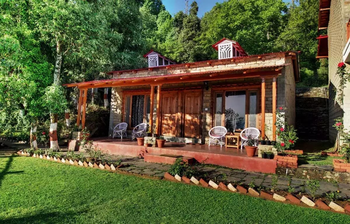 The charming cottage features a wooden facade and a spacious porch, adorned with white wicker chairs and potted plants. Lush greenery surrounds the structure, enhancing the natural setting. Scenic views can be appreciated through the large glass windows, inviting the outdoors inside.