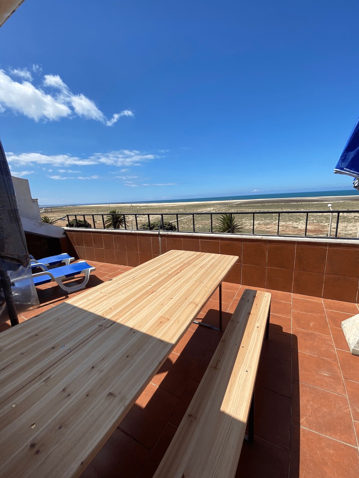 A spacious terrace features a wooden table and benches, providing ample seating for gatherings. The area is bathed in natural light, with a clear view of the ocean from the railing. The landscape includes sandy beach and distant waves under a bright blue sky.