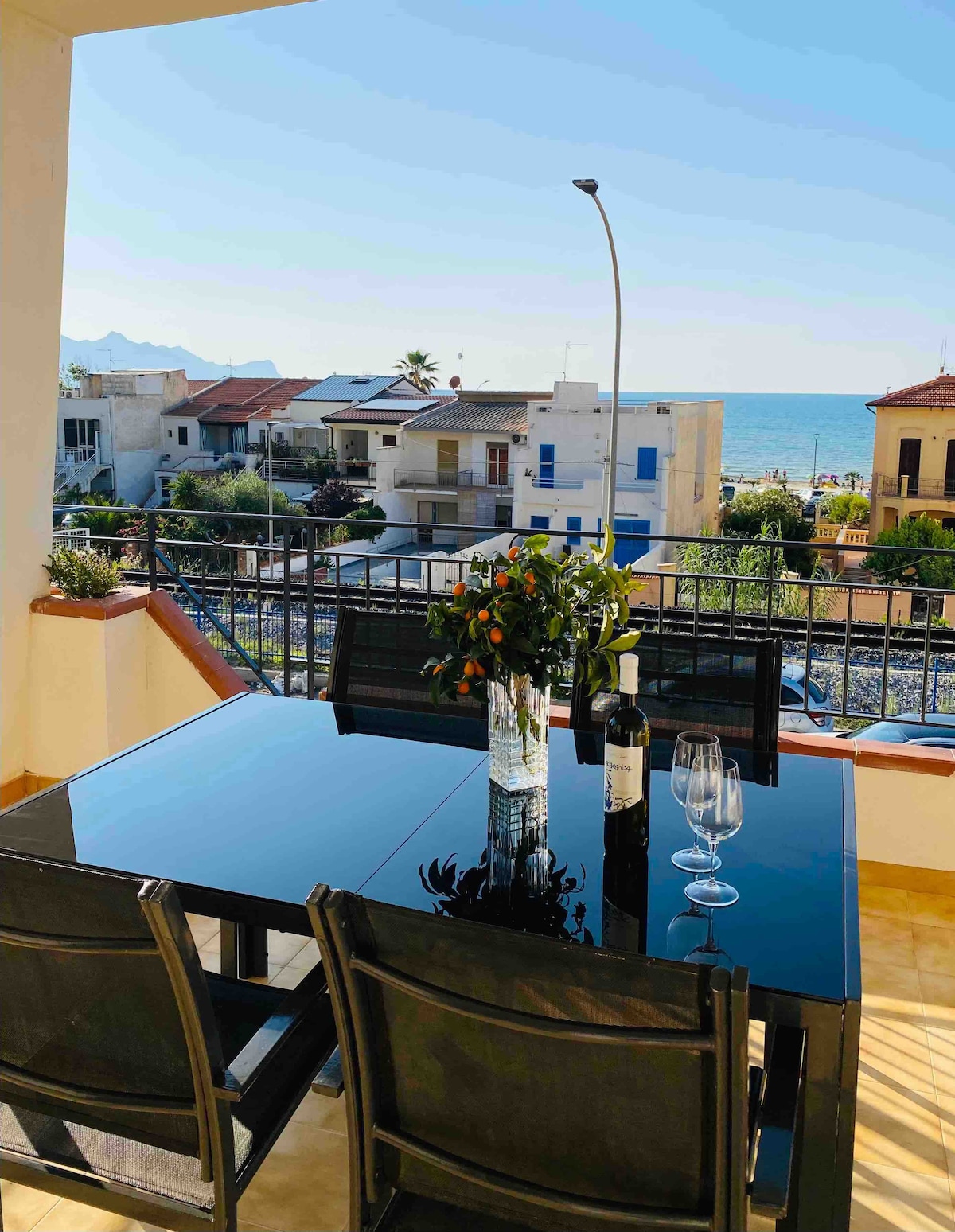 An outdoor veranda is shown with a glass table set for two, featuring a vase of fresh flowers, a bottle of wine, and two glasses. The stunning view of the coastline and the sea is visible in the background, providing a serene environment.