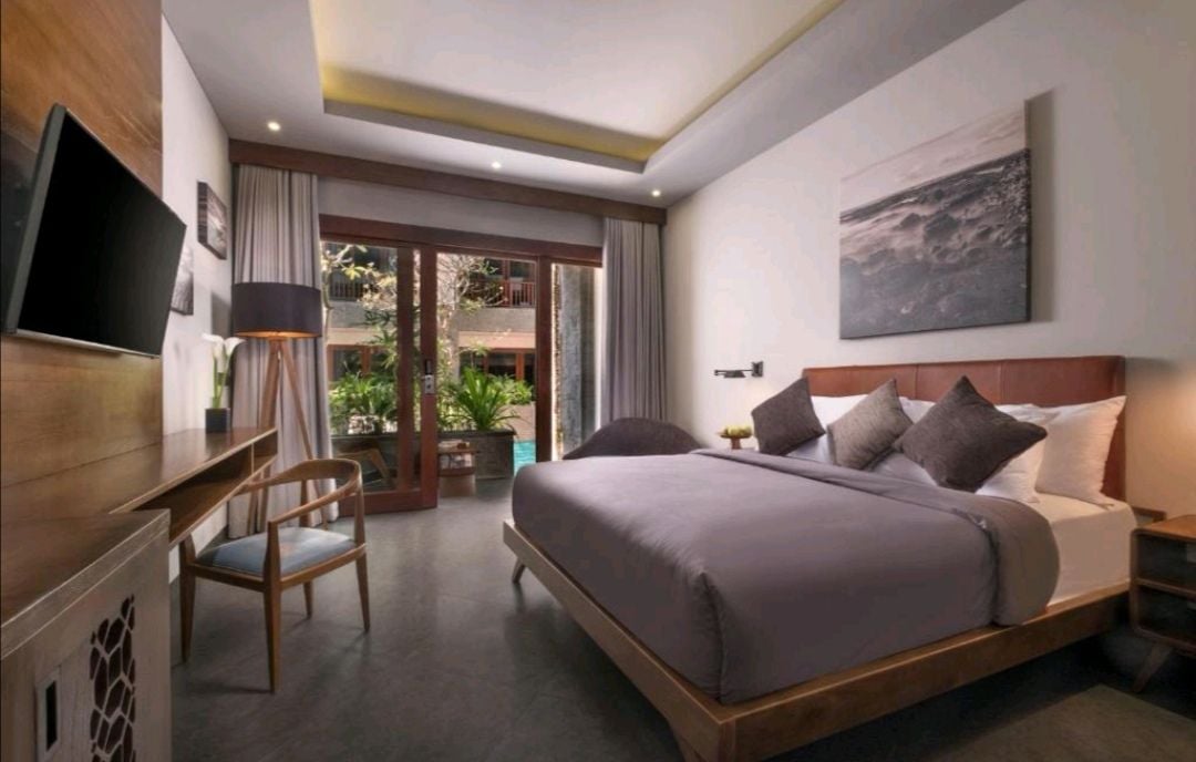 A modern bedroom features a spacious bed with neutral bedding and decorative pillows. Large glass doors provide access to a small outdoor area filled with plants. A wooden desk and chair are positioned nearby, alongside a wall-mounted television.