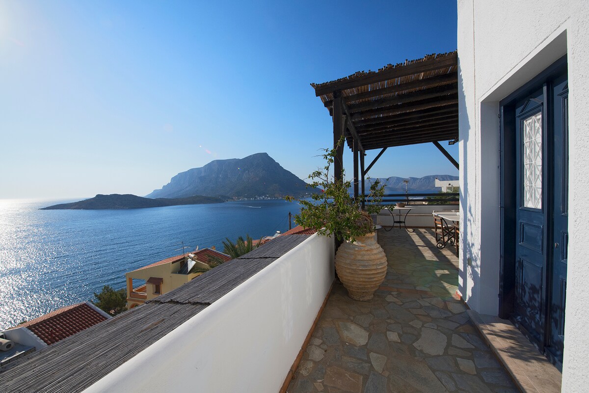 A spacious outdoor terrace is featured, complemented by a wooden pergola. A large stone planter filled with greenery stands beside a seating area, providing views of the shimmering sea and surrounding mountains under a clear blue sky.