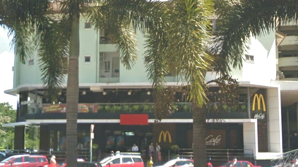 The image shows a ground floor view of a building featuring a McDonald's restaurant, highlighted by its iconic yellow arches. Lush palms frame the scene, with a row of parked vehicles in the foreground. The entrance has outdoor seating with greenery visible in the backdrop.