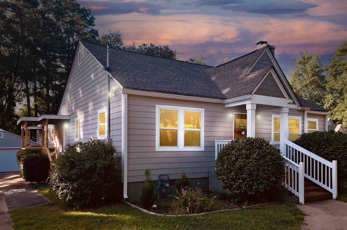 The exterior of a charming bungalow is shown at dusk, with warm lights emanating from the windows. Lush greenery surrounds the home, and a pathway leads toward the welcoming front entrance, creating a serene ambiance in a historic neighborhood.