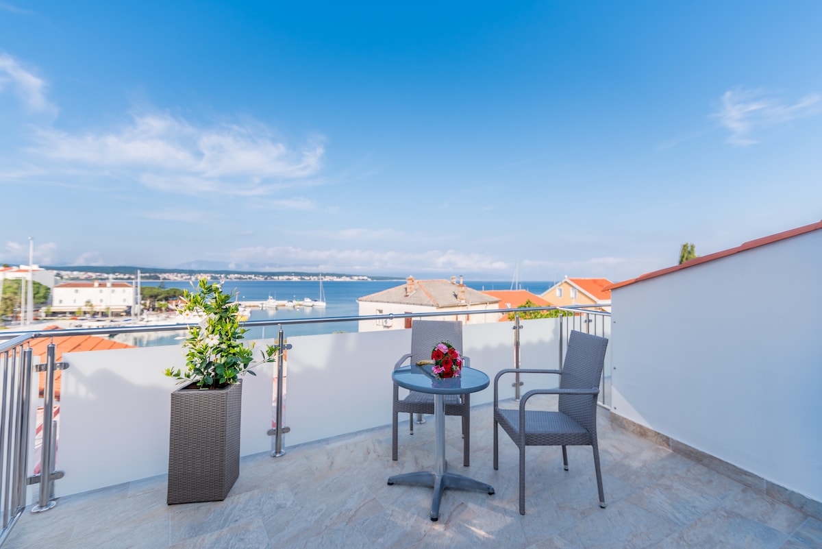 A terrace space featuring a round table and two chairs, complemented by a potted plant. The view of the sea and surrounding rooftops is visible against a clear blue sky, offering a peaceful outdoor setting.
