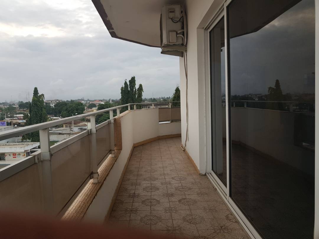 A spacious balcony is shown with a tiled floor and glass railings, providing an unobstructed view of the surrounding area. Floor-to-ceiling glass doors allow natural light into the adjacent indoor space, while greenery is visible in the distance.