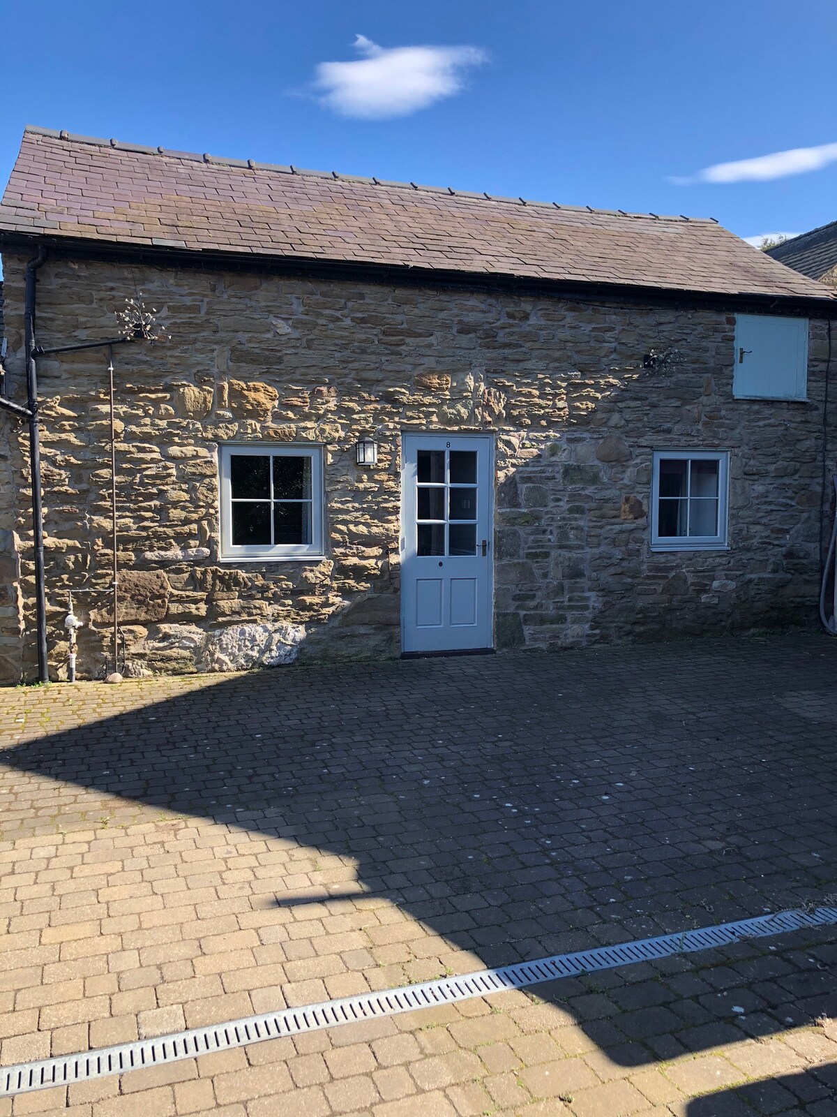 A rustic stone exterior of the barn is visible, featuring a wooden door and two windows. Sunlight casts shadows on the patterned pavers of the courtyard, which offers a peaceful entrance to the retreat.