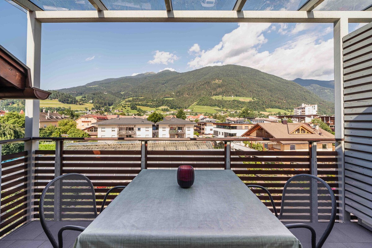 The large terrace features a table with outdoor seating and offers expansive views of the surrounding mountains and lush greenery. The structure includes a glass roof for shade while allowing natural light to brighten the space.