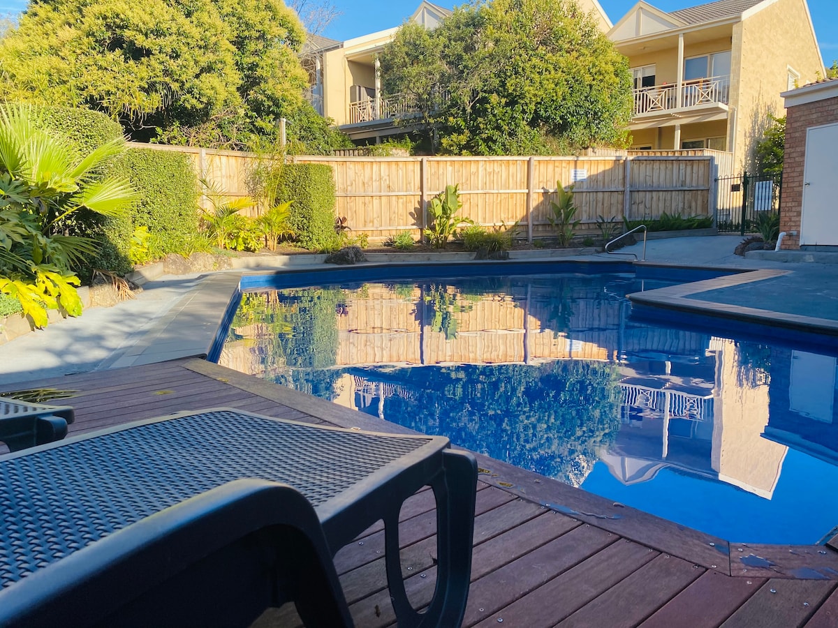 A shared outdoor pool is surrounded by lush greenery and wooden decking. The pool's clear blue water reflects the blue sky, and lounge chairs are positioned nearby, providing a comfortable area to relax and enjoy the serene garden setting.