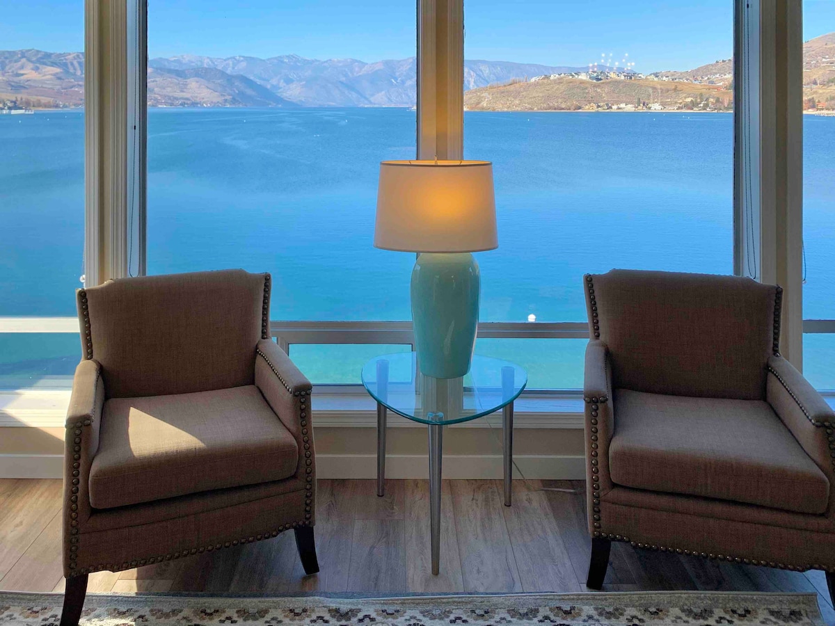 Two comfortable armchairs are positioned by a large window, offering unobstructed views of the serene lake and mountains. A small glass table rests between them, topped with a softly glowing lamp. Natural light fills the space, enhancing the calm ambiance.