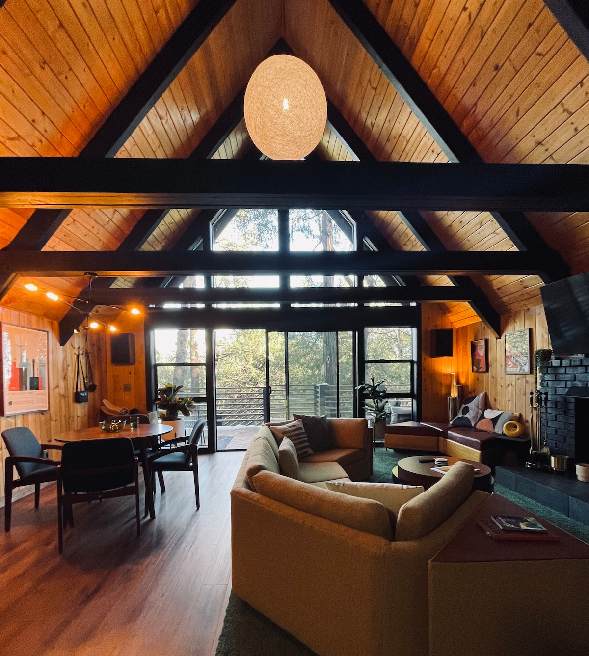 The interior space showcases a blend of wooden accents and natural light, with vaulted ceilings and large windows that frame forest views. Comfortable seating areas and a dining table are arranged to encourage gathering. A modern light fixture adds a contemporary touch to the cozy atmosphere.