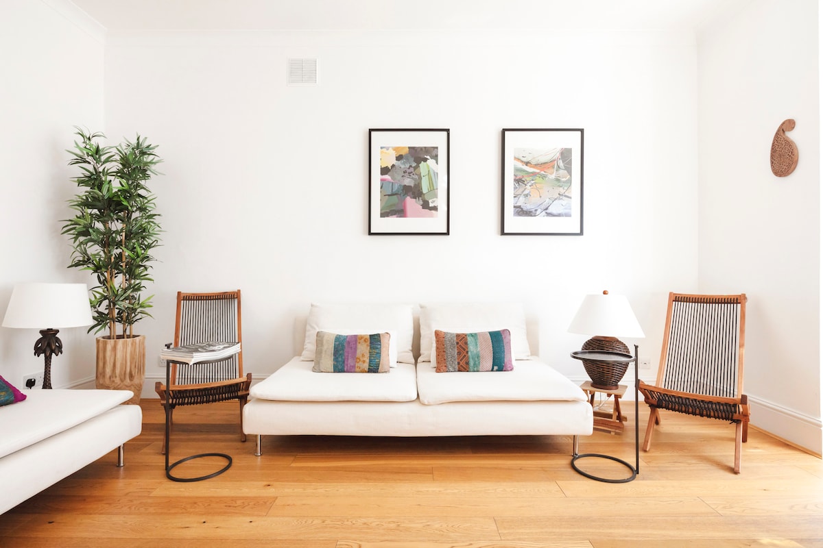 A light-filled living area features a white sofa adorned with colorful cushions. Two stylish chairs flank the sofa, while a small, round table and table lamps provide functionality. Framed artwork adds a touch of personality to the minimalist walls.