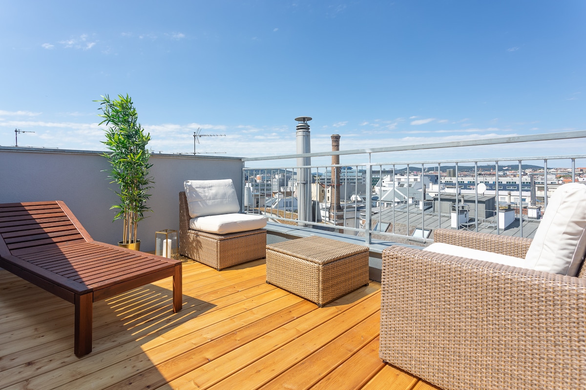 A private terrace features wooden flooring, showcasing a sun lounger and a stylish seating arrangement with wicker furniture. Plants add a touch of greenery, while the railing offers unobstructed views of the surrounding rooftops and skyline under a clear blue sky.