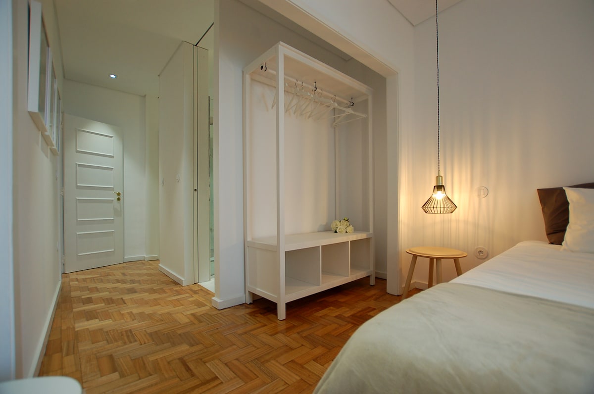 A bright sleeping area features a spacious bed with soft linens, complemented by a light wooden side table. A hanging light fixture provides warm illumination, and a minimalist wardrobe is positioned nearby. The room is designed with neutral tones and natural light is visible from the hallway.