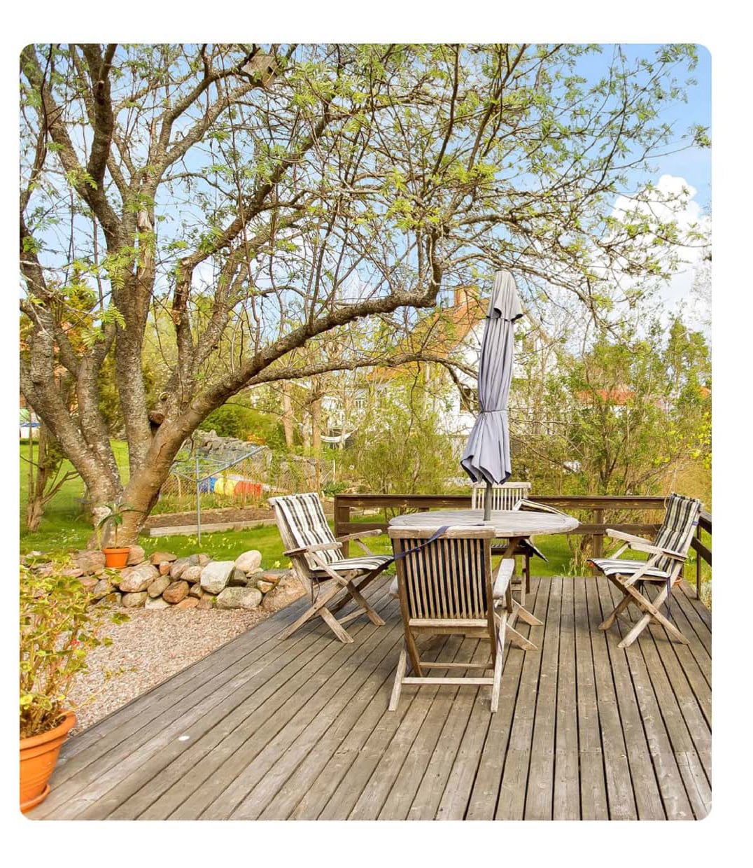 A spacious outdoor deck is furnished with a dining table and chairs, shaded by a tall umbrella. A sturdy tree provides natural cover, while a grassy area with a trampoline is visible in the background. Potted plants add greenery to the setting.